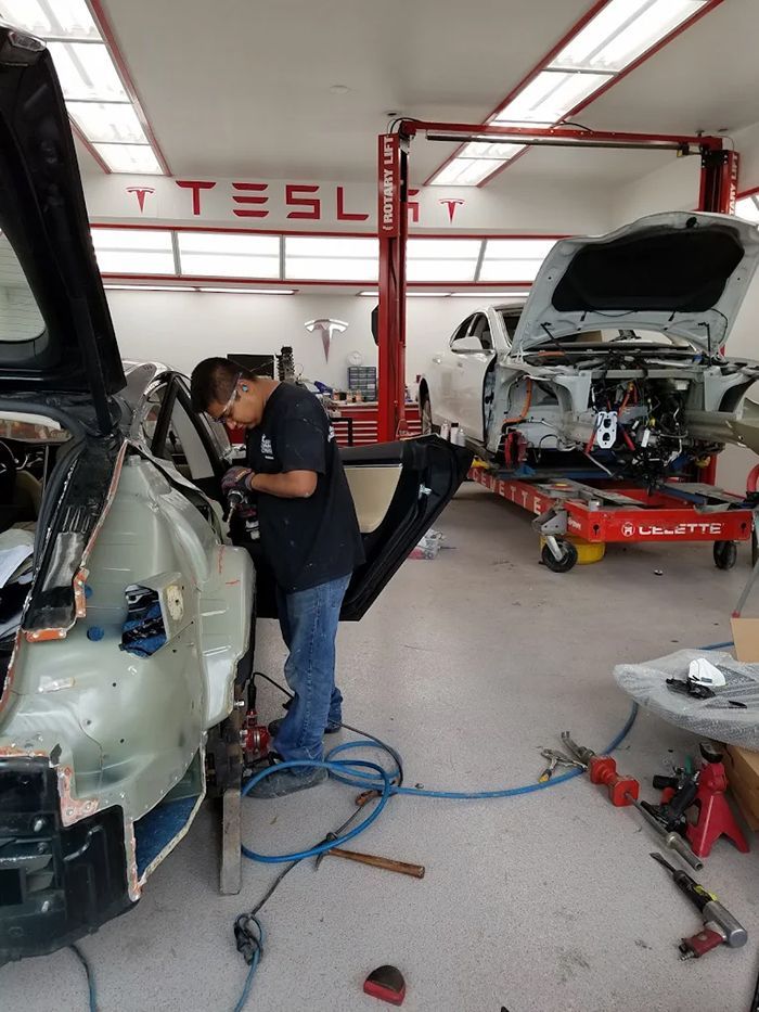 Man repairing a Tesla in a workshop. Car body panels are removed. The Tesla logo is visible.