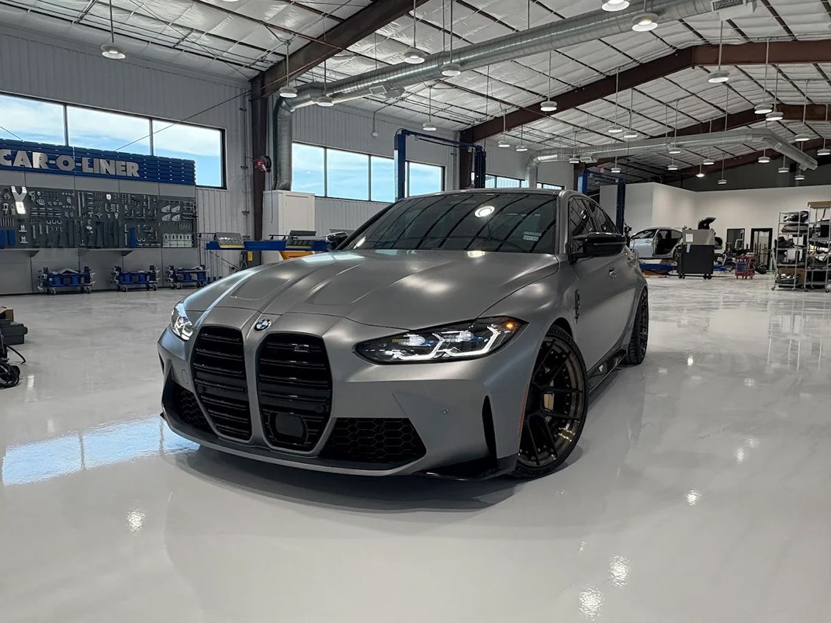 Gray BMW M3 sedan in a well-lit garage, with a glossy floor and industrial setting.