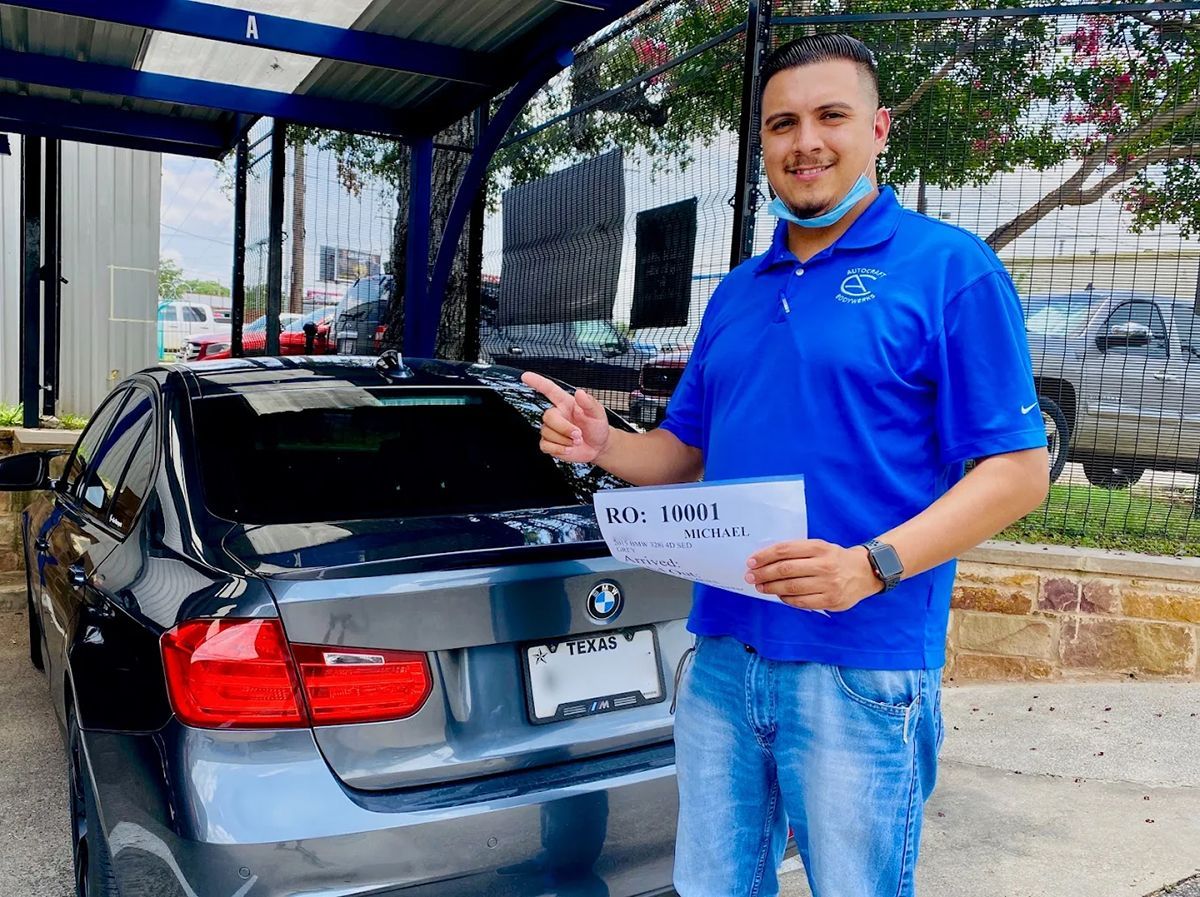 Man points at a gray BMW, holding a sign. He wears a blue shirt and jeans, standing at a carwash.
