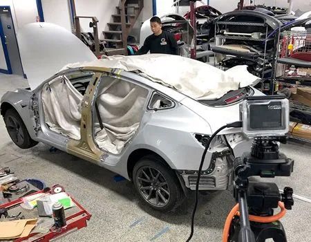 A person works in an auto body shop on a silver car stripped of its doors and trunk, with a camera filming on a tripod.