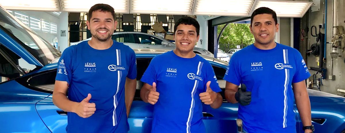 Three men in blue shirts give thumbs-up near a blue car in a garage.