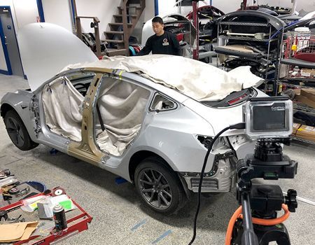A man repairs a silver Tesla in a workshop. Car is partially disassembled. A camera is set up.