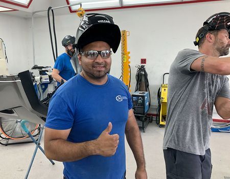 Man in blue shirt, welding helmet, giving thumbs up in a workshop; two others weld in the background.
