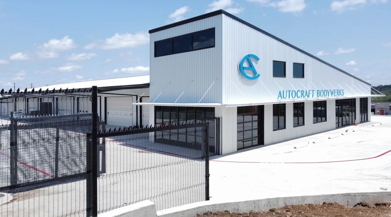 Exterior of Autocraft Bodywerks building spicewood tx with a blue logo on a white wall. Black windows. Sunny day.