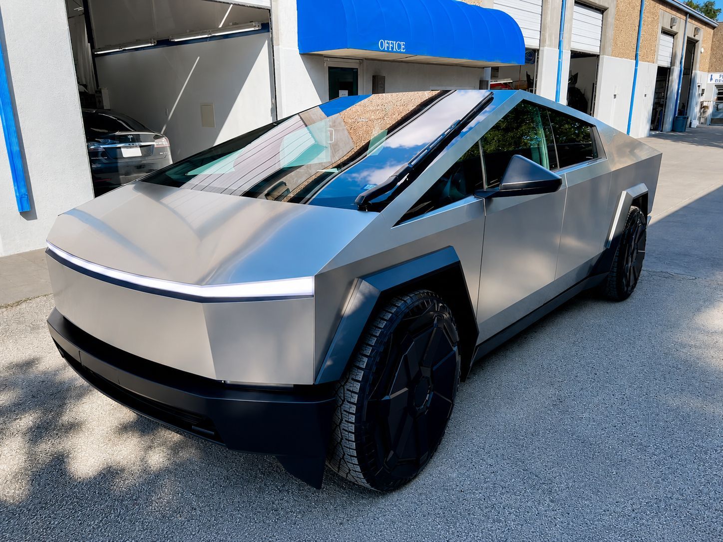 Tesla Cybertruck, angular silver-gray body, black wheels, parked outdoors in front of a building.