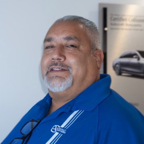 Man in a blue polo shirt standing indoors, smiling slightly, with a car poster in the background
