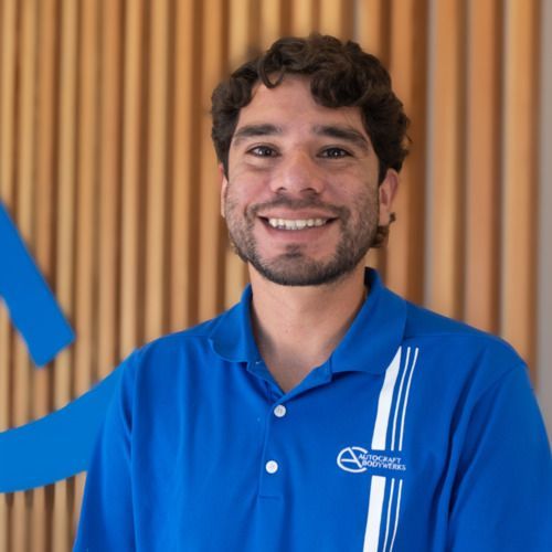 Man in a blue polo shirt standing indoors, smiling slightly, with a car poster in the background