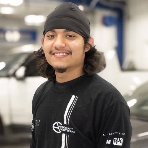 Person smiling in a black beanie and shirt, standing in a garage with blurred cars in the background