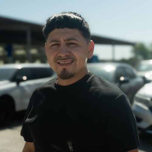 Man in a black shirt standing outdoors in bright sunlight with parked cars behind him