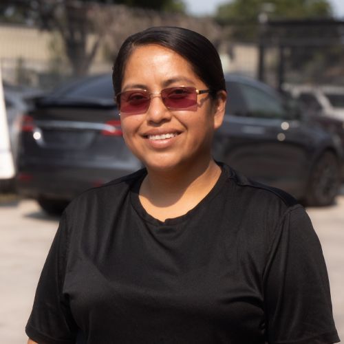 Smiling person in a black shirt and tinted glasses standing outdoors with cars in the background