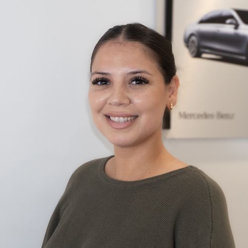 Smiling woman in an olive sweater standing in a showroom with a car poster behind her