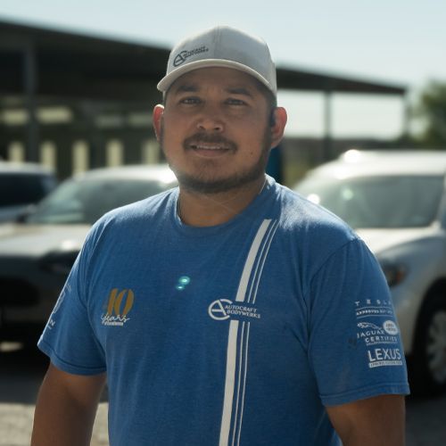 Man in a blue shirt and cap standing outdoors in front of parked cars and a building