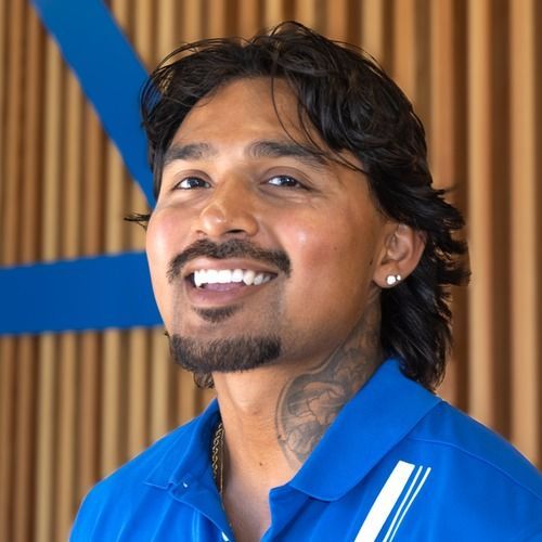 Smiling man in a blue shirt with a neck tattoo, standing in front of wood slats and blue accents