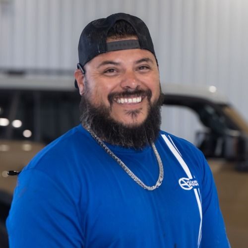 Smiling man in a blue shirt and black cap standing indoors beside a vehicle