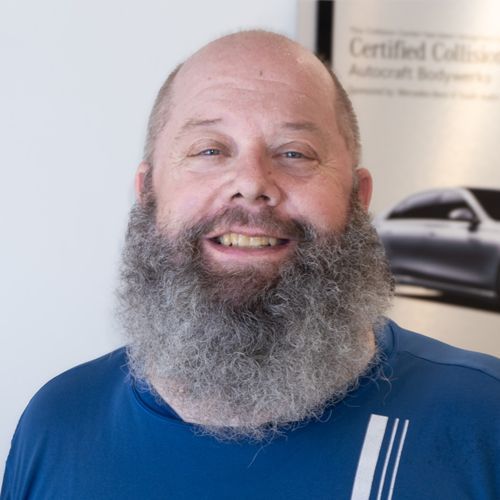 Smiling bearded man in a blue shirt, seated indoors with a car poster behind him