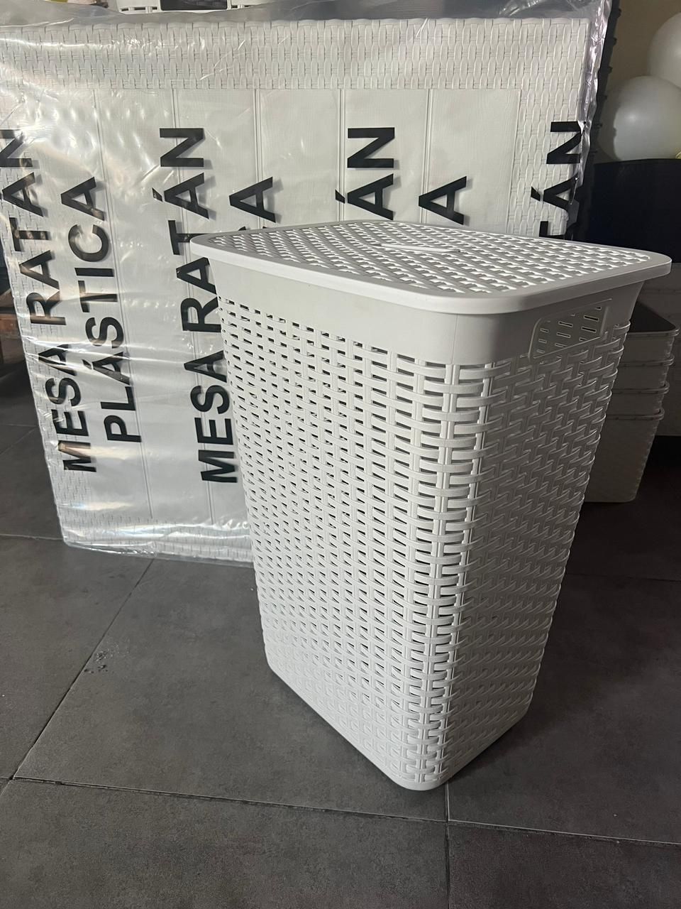 Cesta de ropa tejida de color marrón con tapa sobre un suelo de baldosas junto a sacos etiquetados como “MESA RATA”.