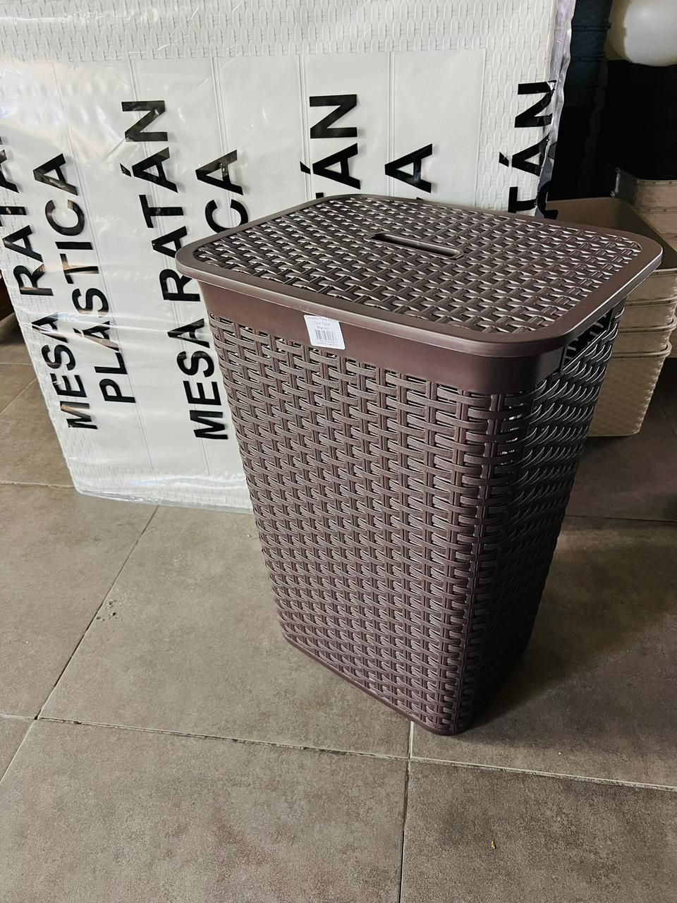 Cesta de ropa tejida de color marrón con tapa sobre un suelo de baldosas junto a sacos etiquetados como “MESA RATA”.