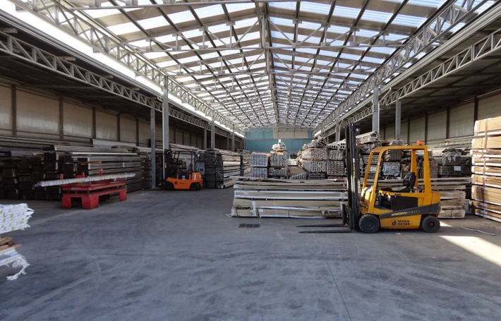Ampio interno di un magazzino con scaffalature metalliche, materiali impilati e un carrello elevatore giallo su un pavimento di cemento.