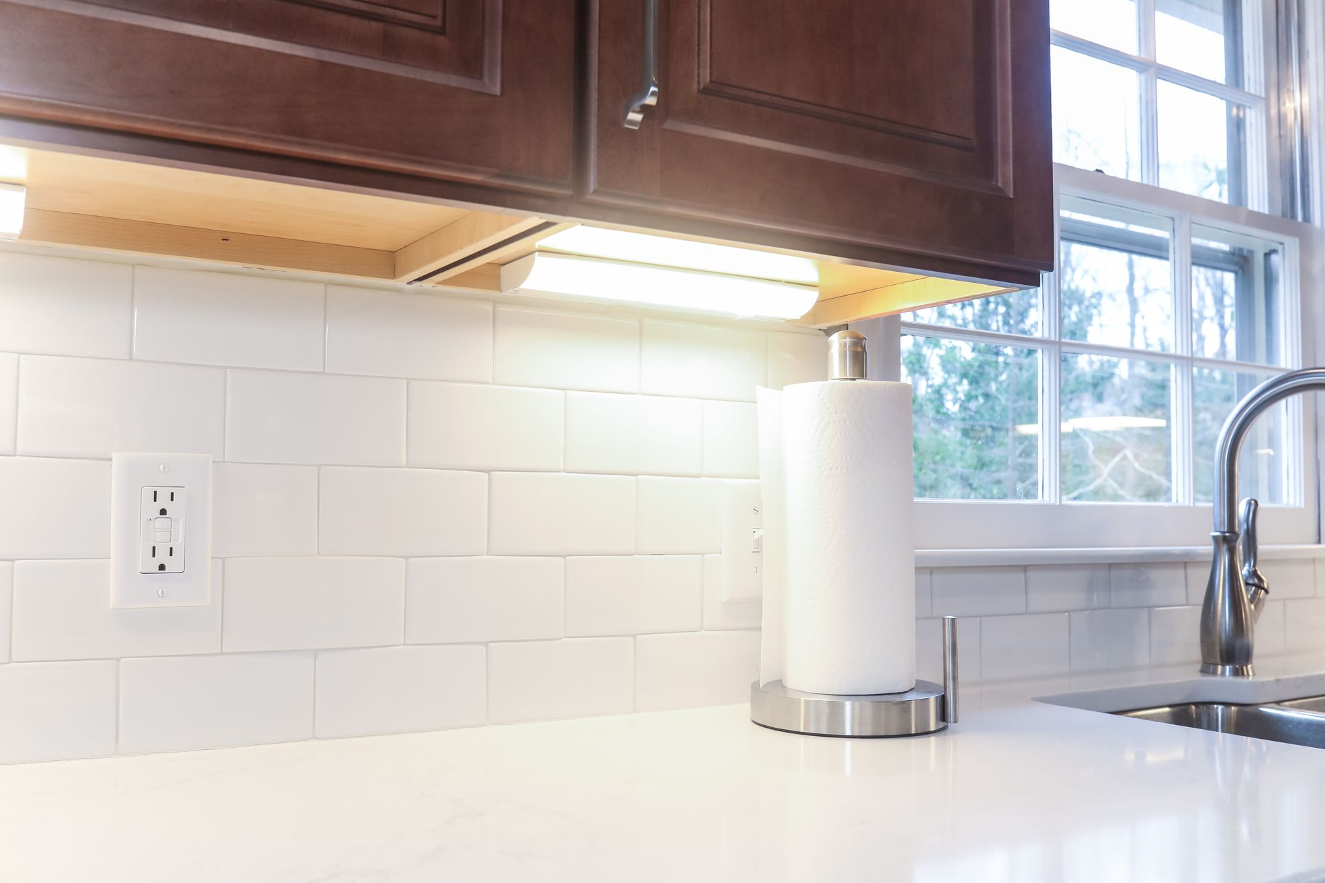 Modern kitchen countertop with white tile backsplash, wood cabinets, paper towel roll, and sink by a window