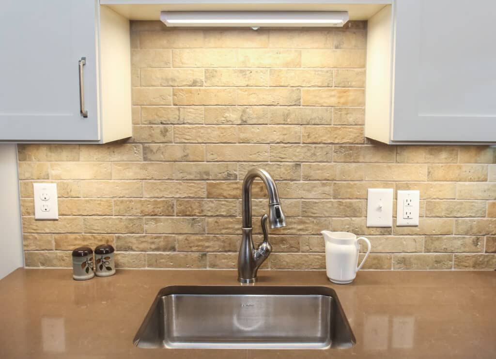 Kitchen sink with stainless faucet under cabinet lights, beige brick backsplash, and brown countertop