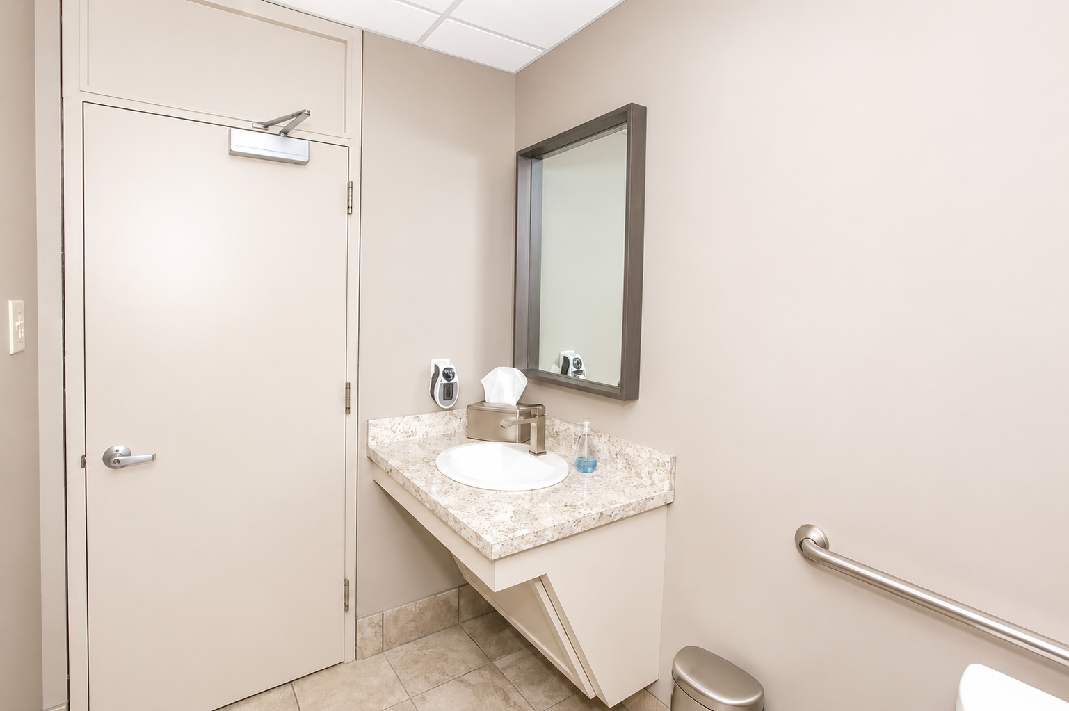 Accessible restroom with sink, mirror, door, and grab bar in a beige-tiled interior