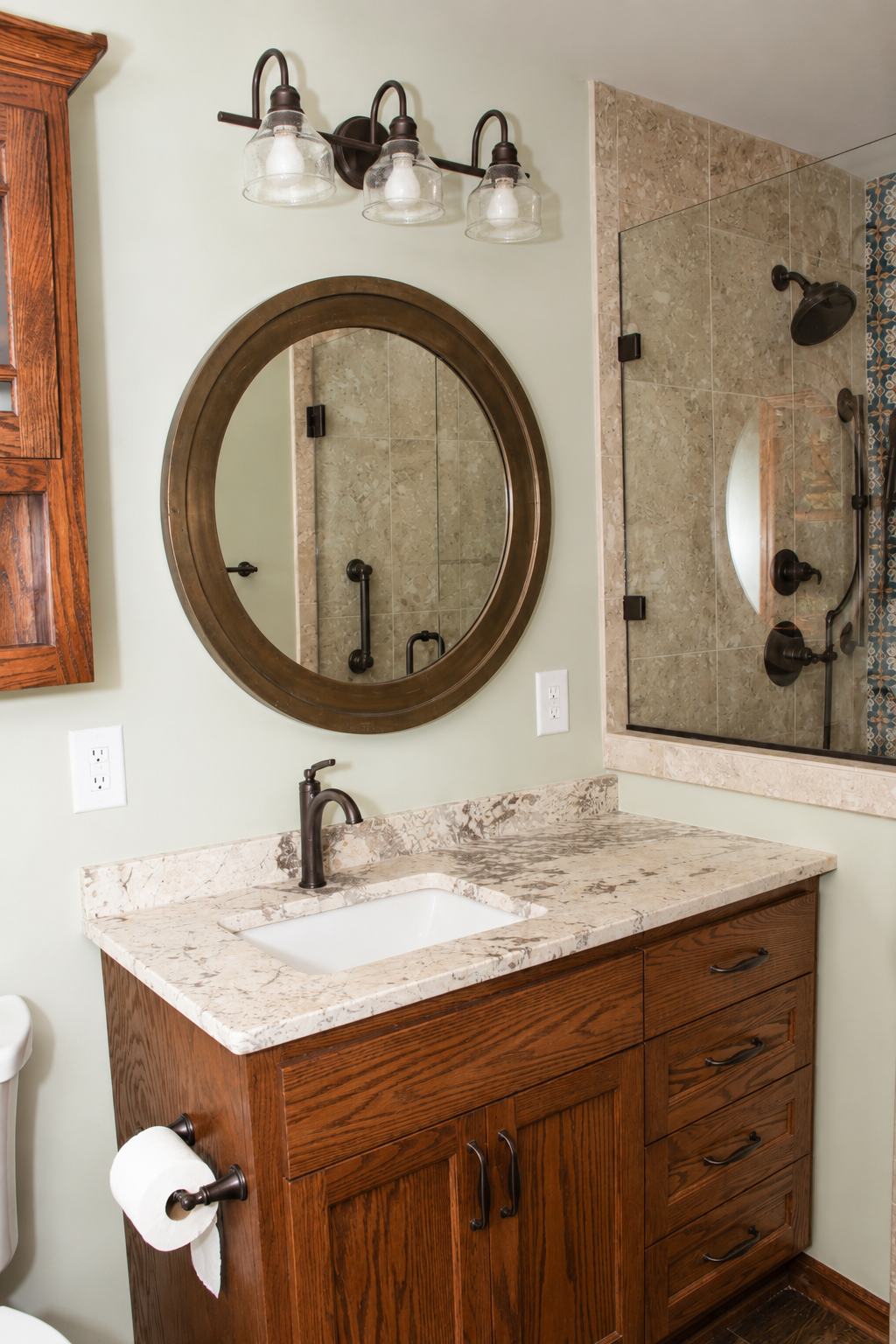 Bathroom vanity with wood cabinet, round mirror, granite countertop, and glass shower door