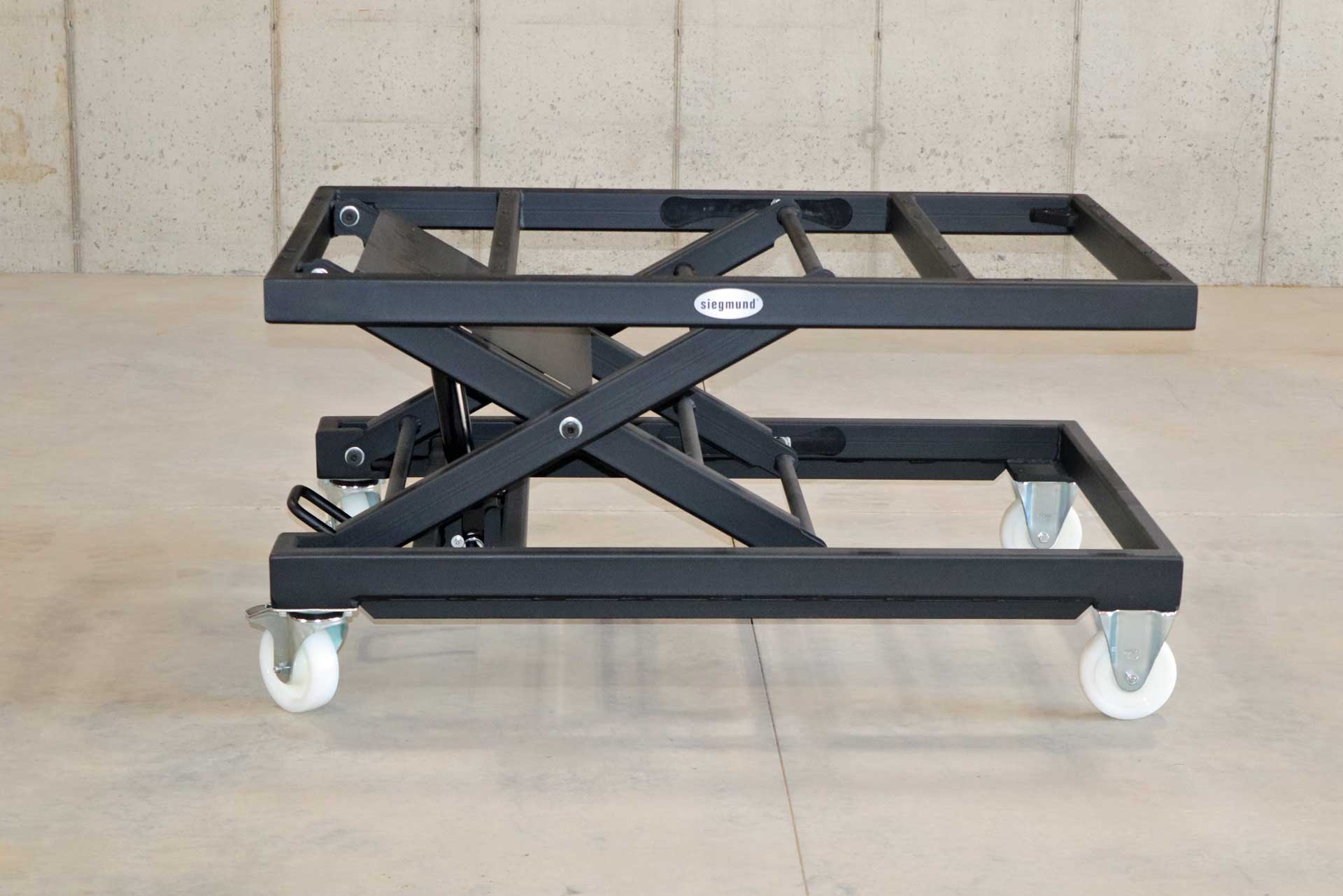 Siegmund Lifting Table A black scissor lift with white wheels is sitting on a concrete floor.