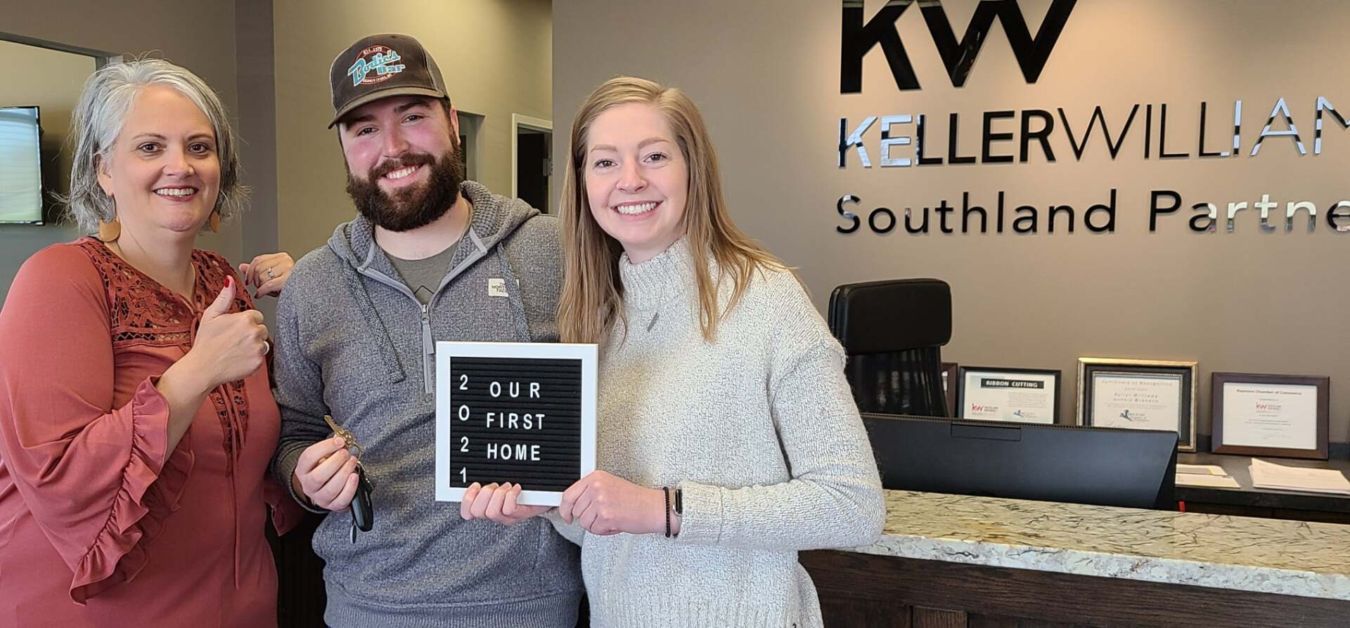 Three people in a Keller Williams office celebrating a home purchase. One person holds a sign that reads 