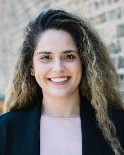 Woman with long, wavy hair smiles, wearing a black blazer and light pink top, against a brick wall.