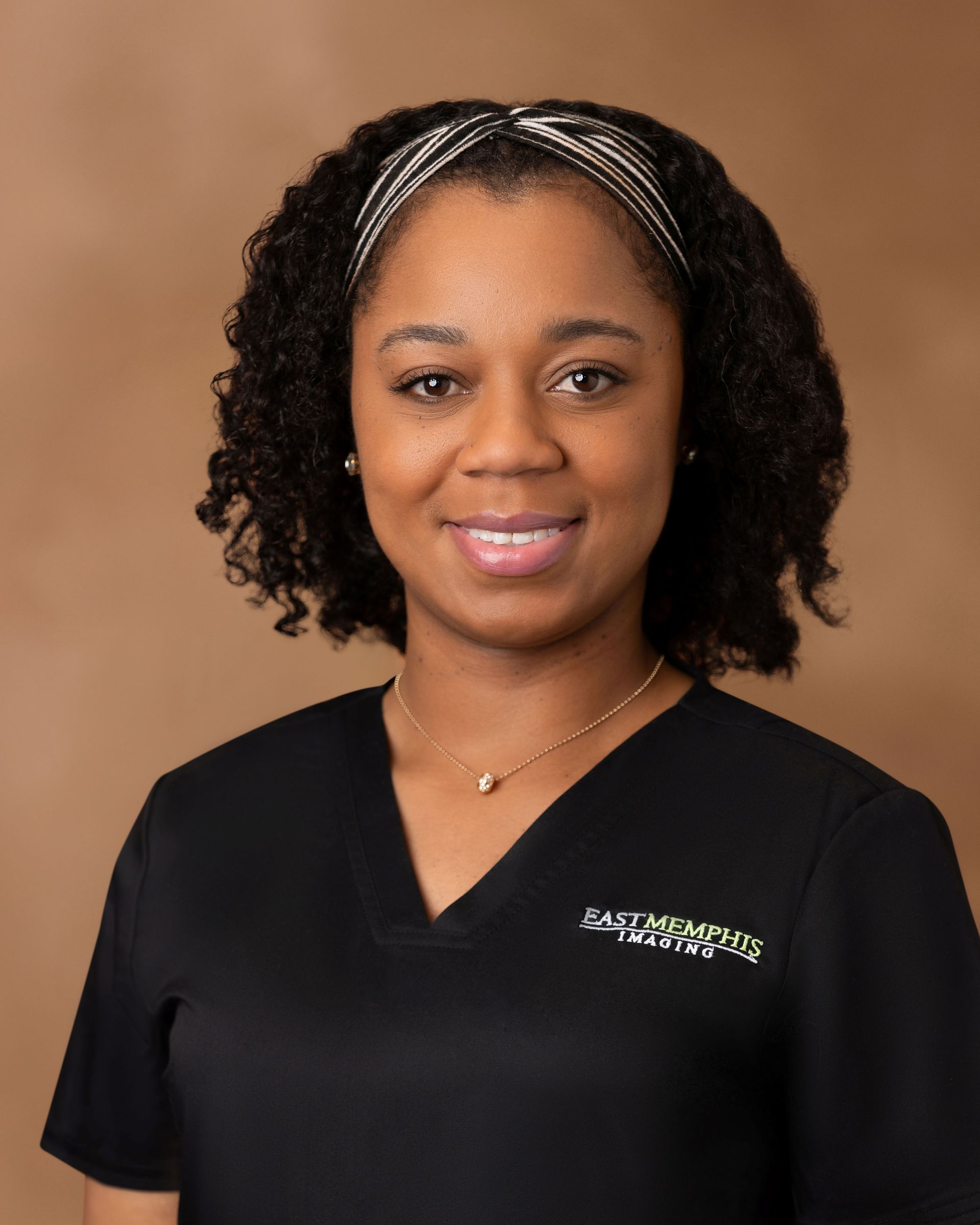 Smiling person in black top against a brown studio backdrop, wearing a silver headband and necklace.