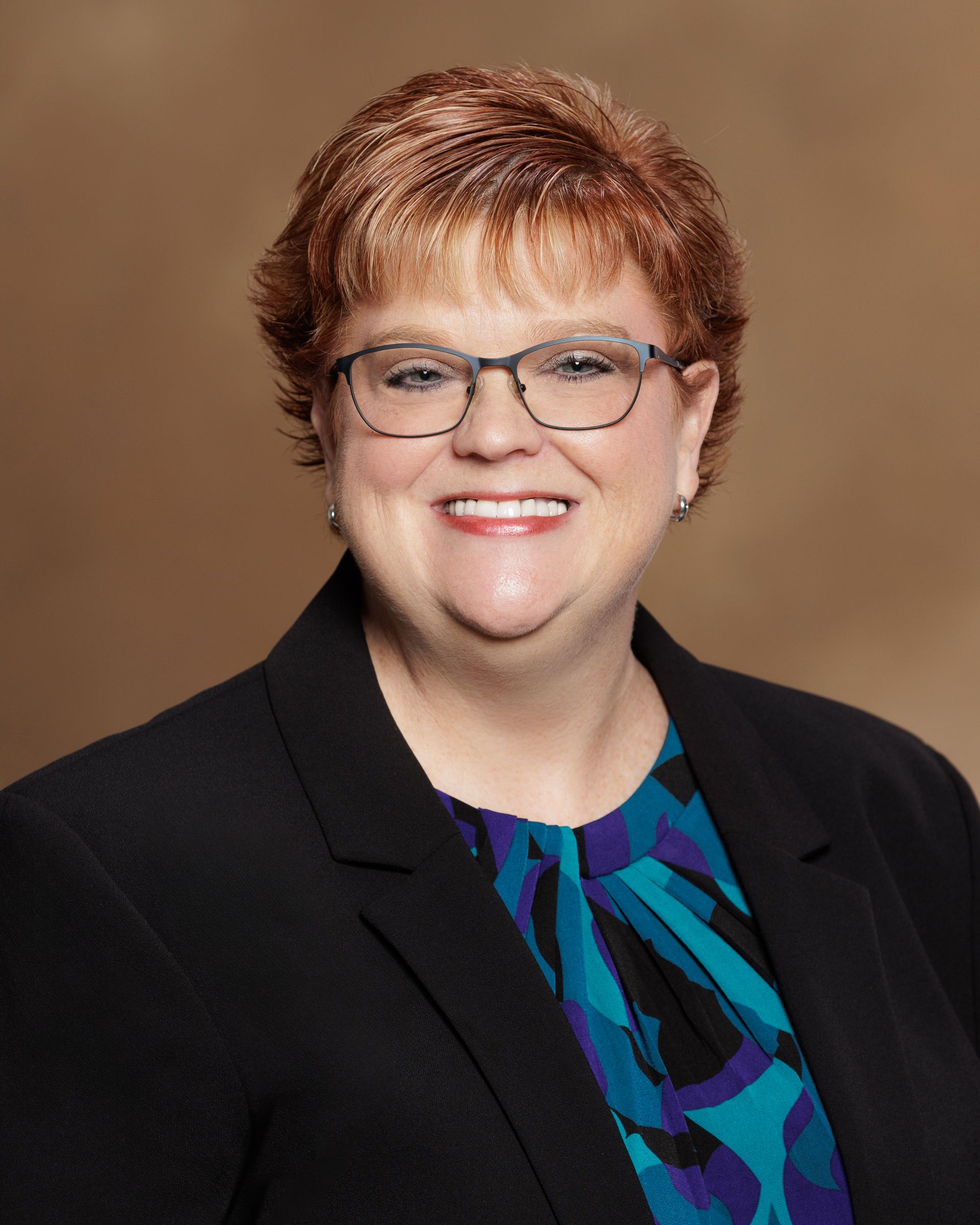 Smiling professional in a black blazer and blue patterned top against a brown studio background