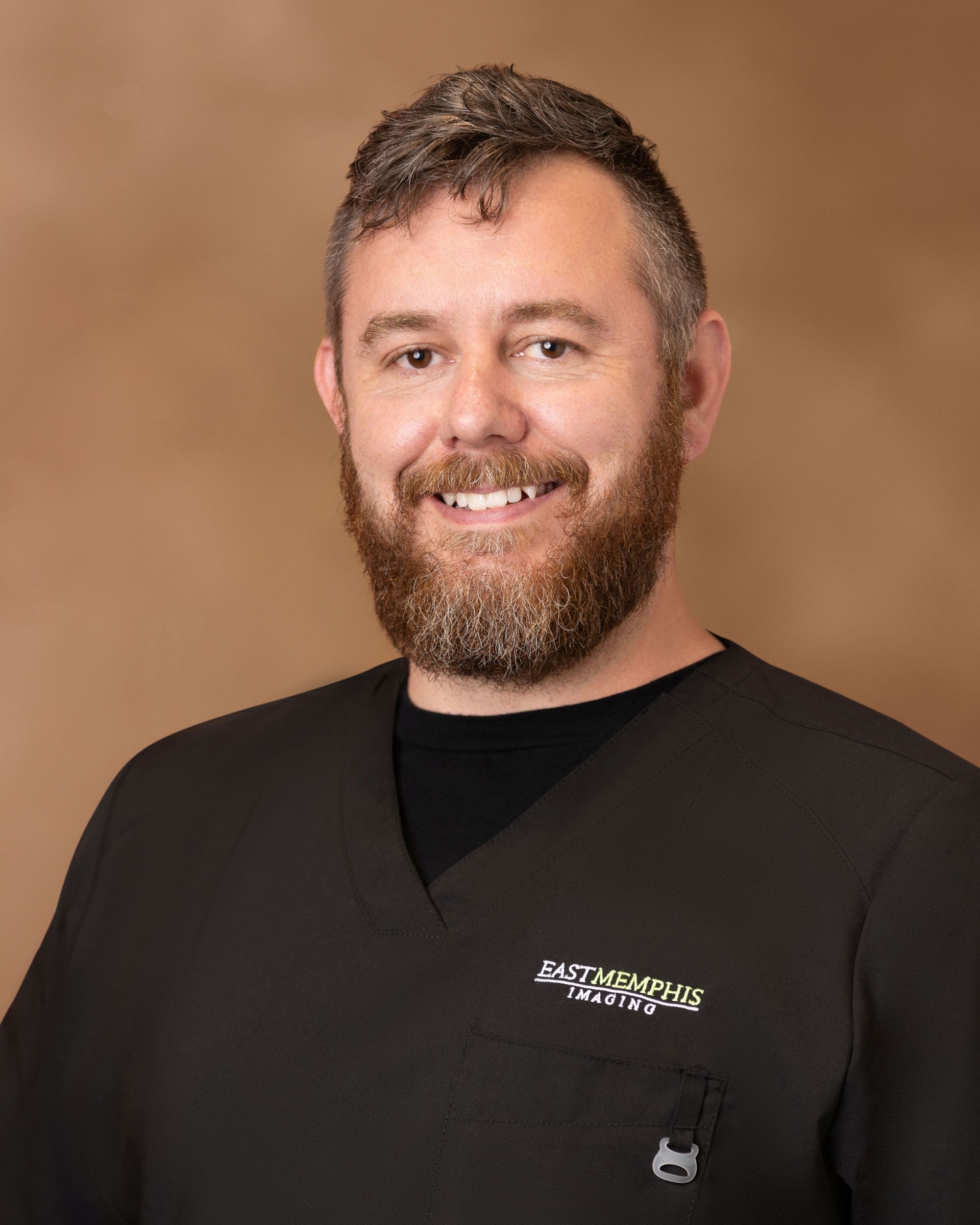 Smiling person in a black shirt with a logo, standing against a brown background.