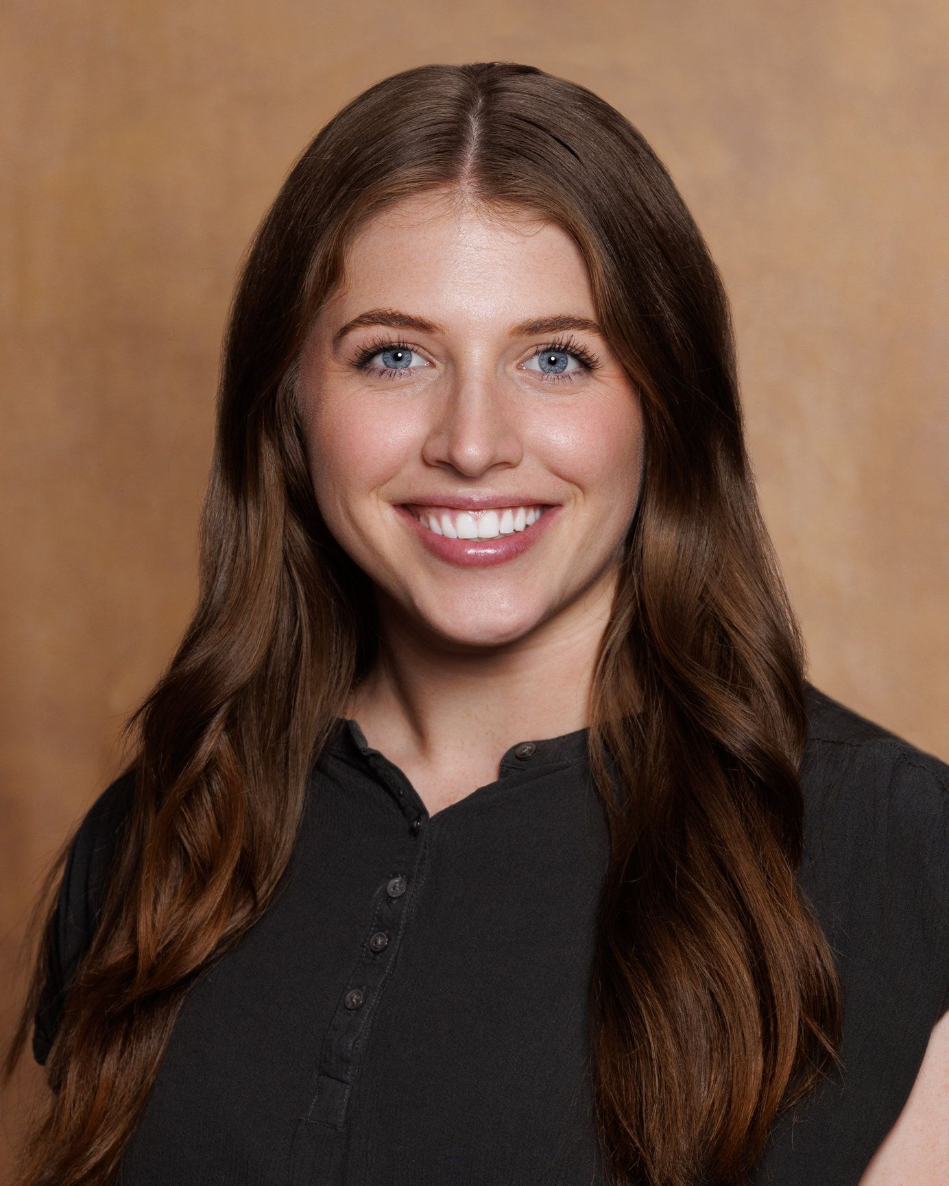 Portrait of a smiling person with long brown hair wearing a black top against a tan background