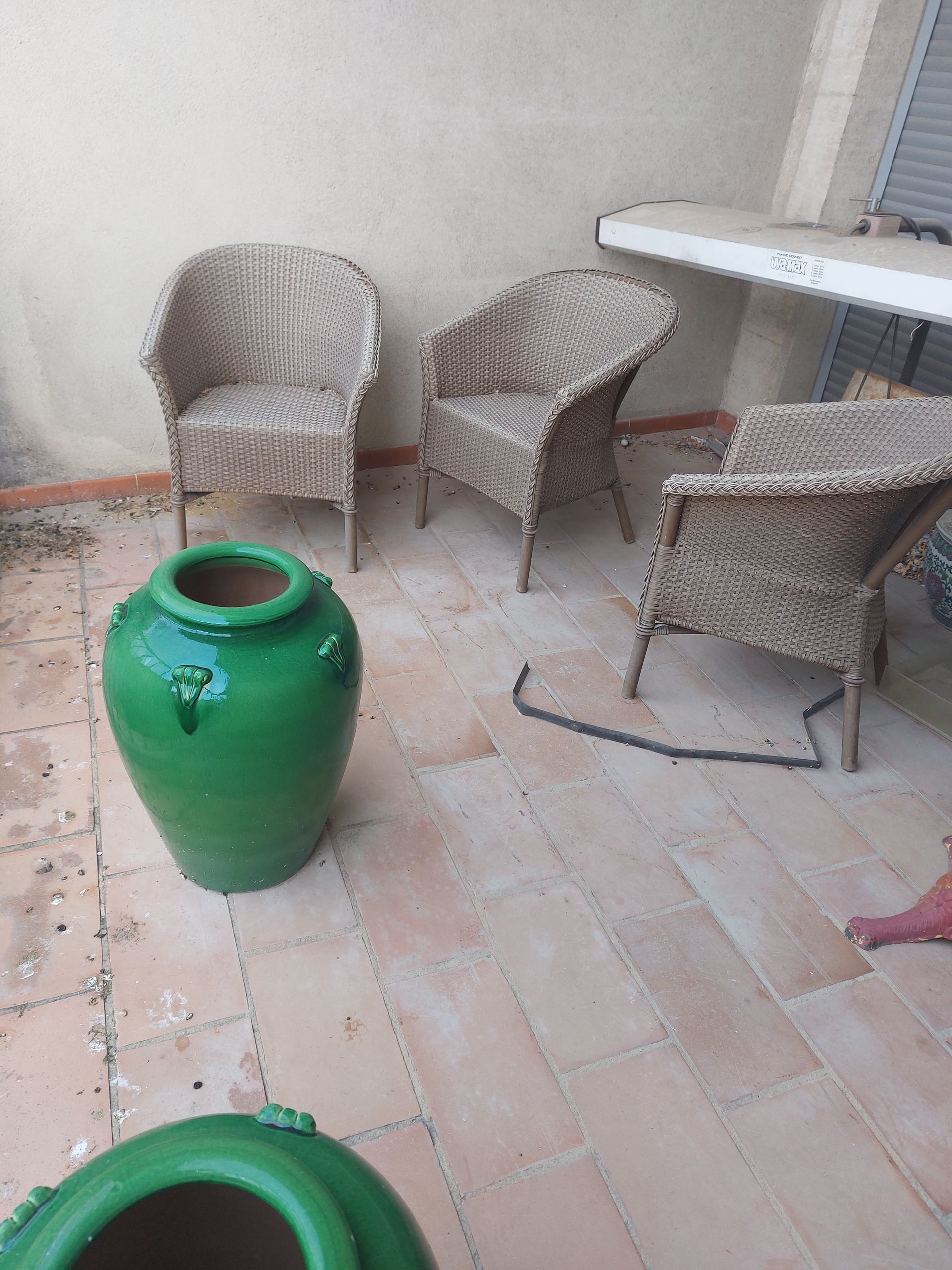 Un vase vert est posé sur un sol carrelé à côté de chaises en osier.