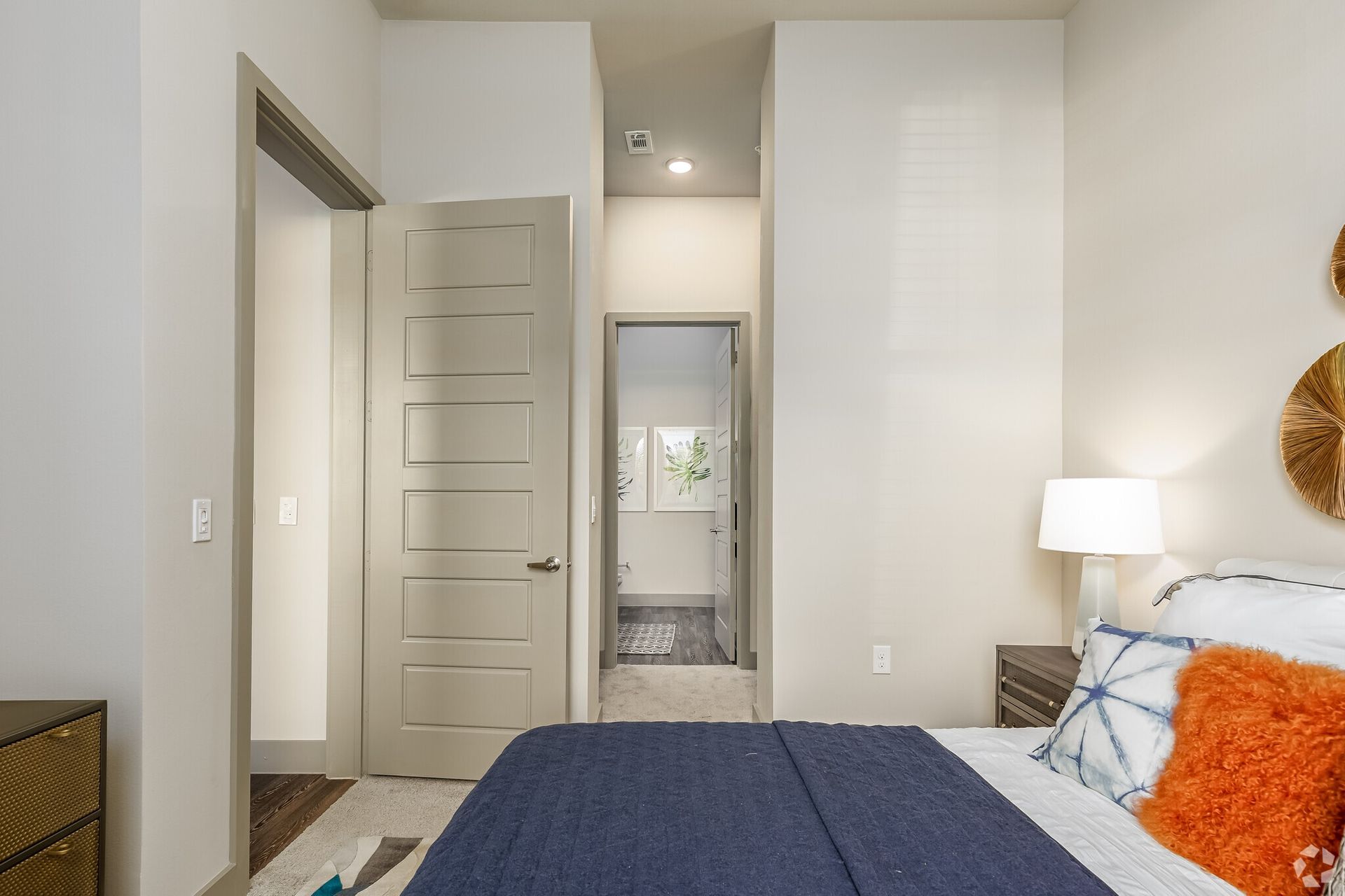 Bedroom in a modern apartment with a bed, nightstand, lamp, and doorway to a hallway.
