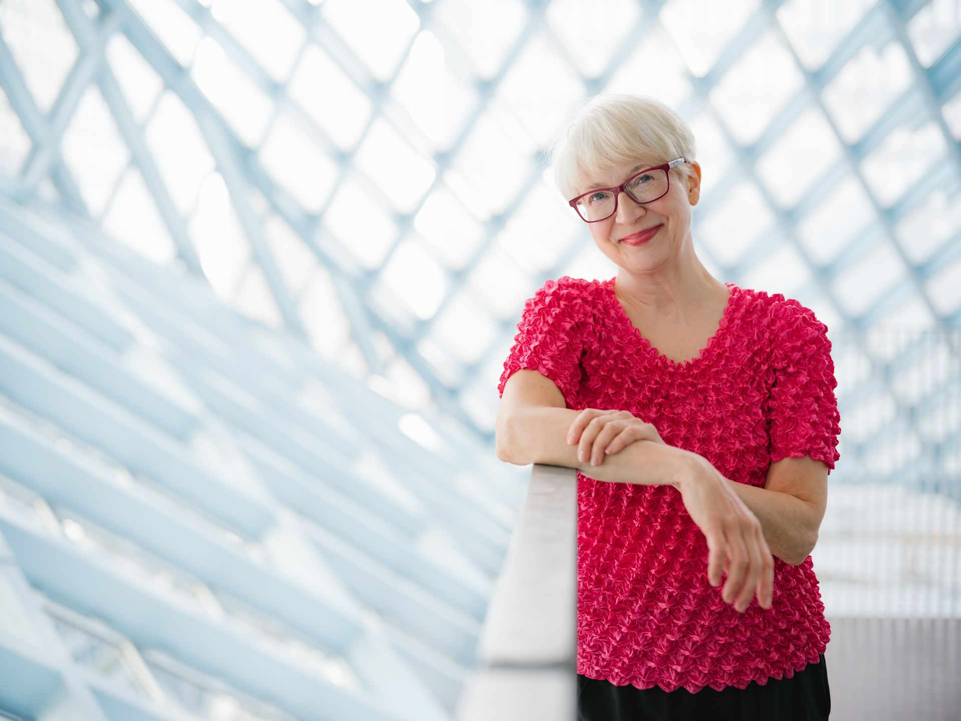 Andrea wears glasses and a pink top and is leaning on a railing.