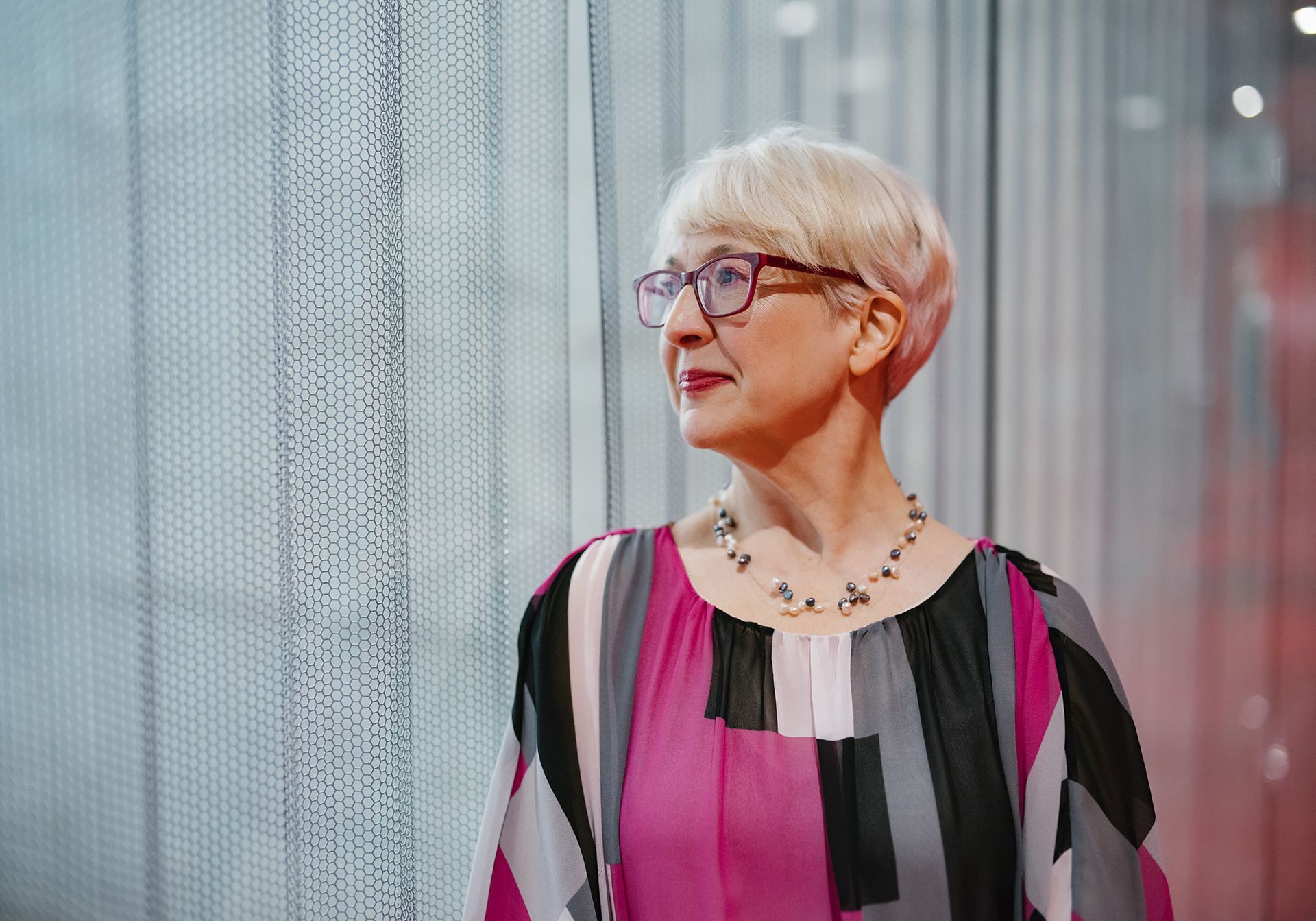 Andrea is wearing glasses and a necklace standing in front of a window.