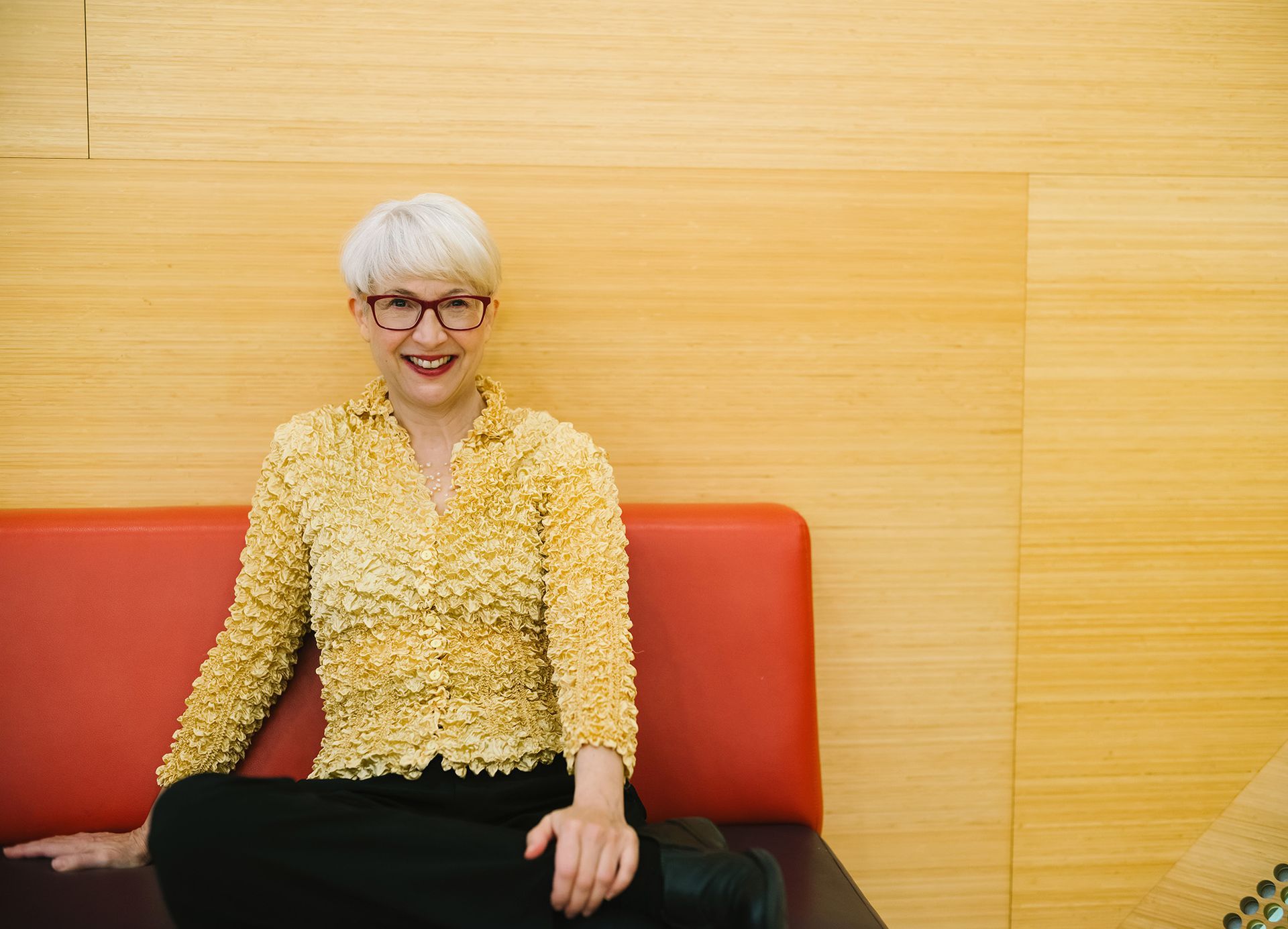 Andrea is sitting on a red couch wearing glasses and a yellow shirt.
