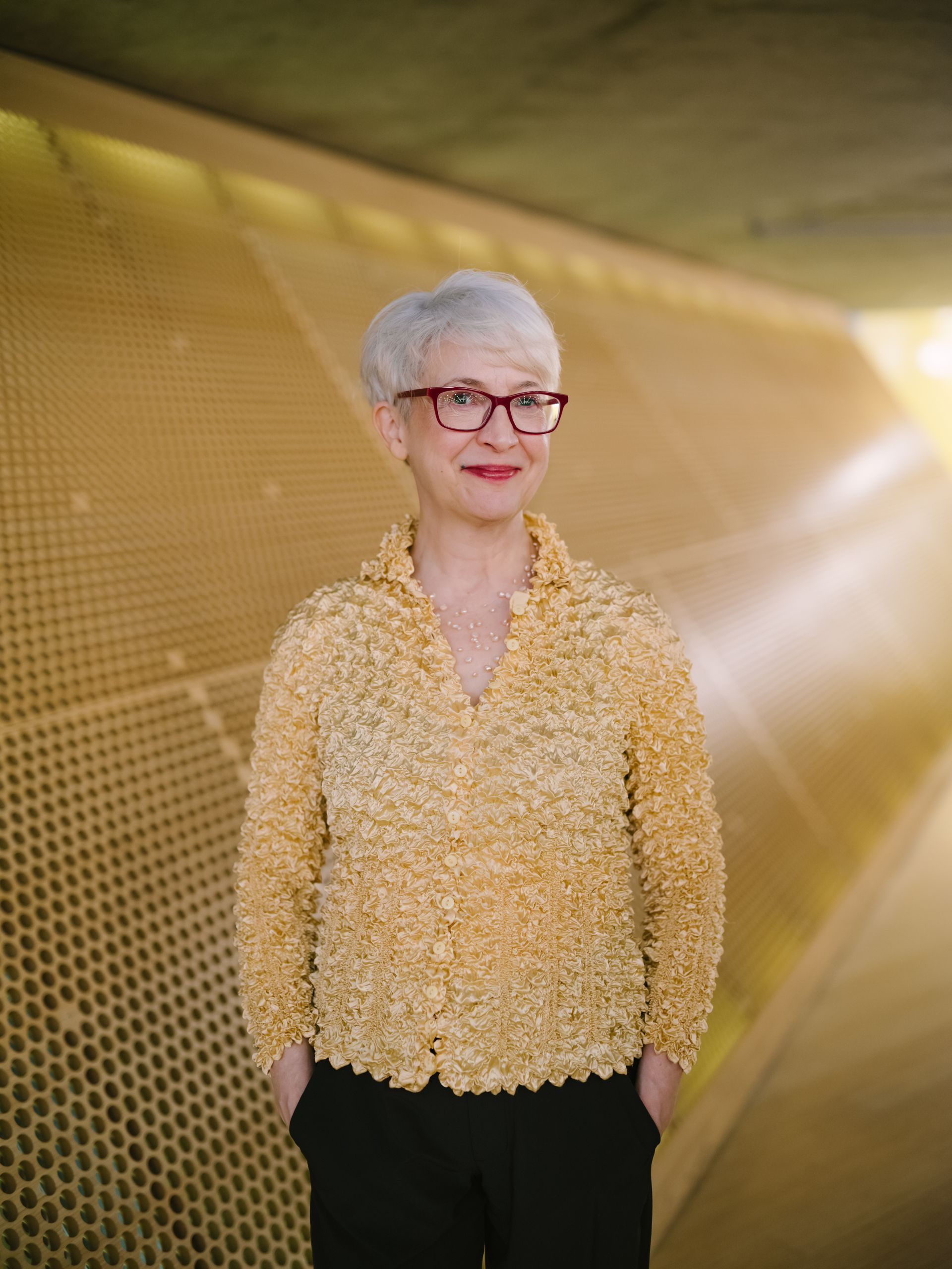 Andrea wears glasses and a yellow shirt and is standing in front of a gold wall.