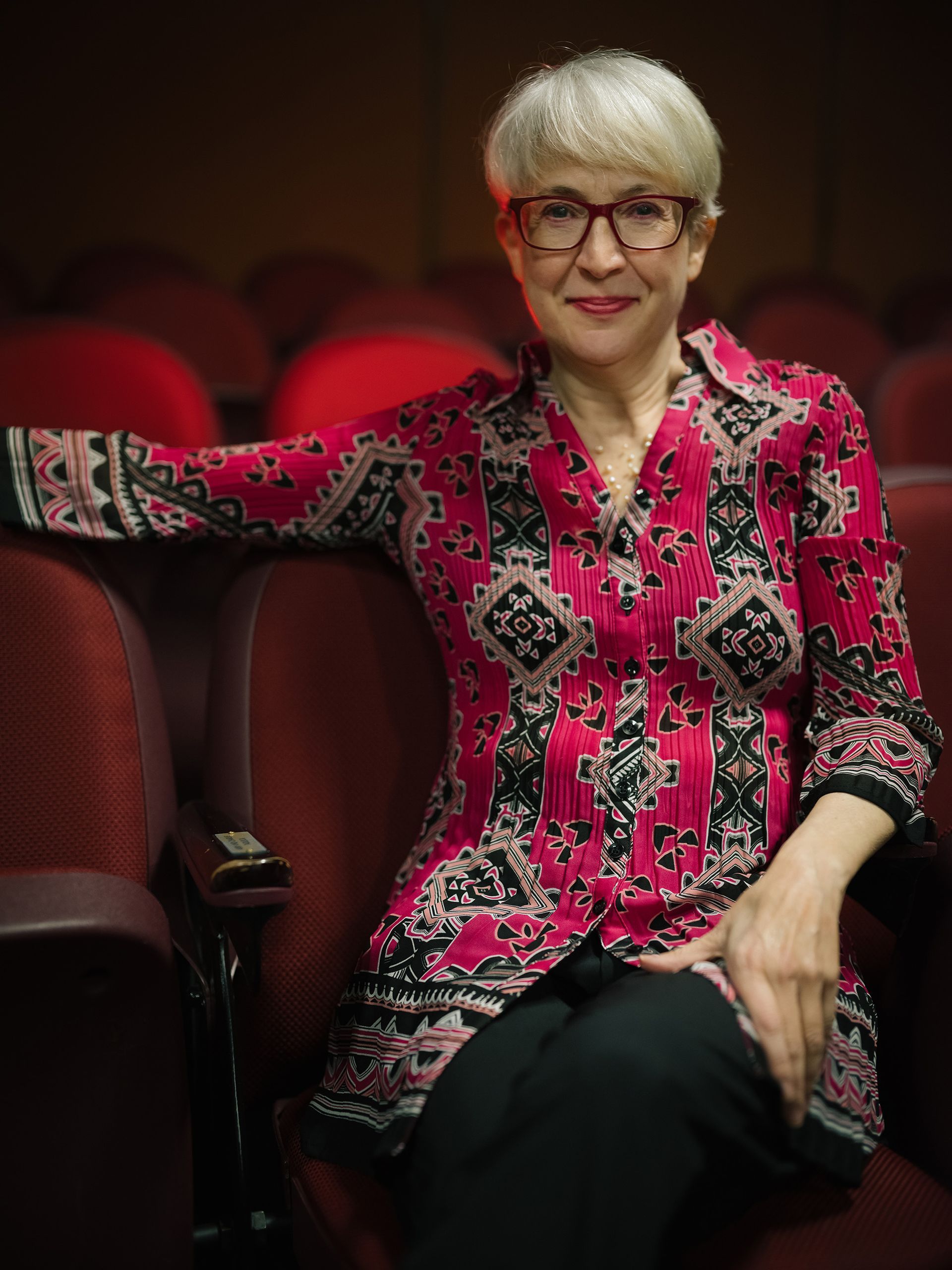 Andrea in a pink shirt and glasses is sitting in an auditorium.