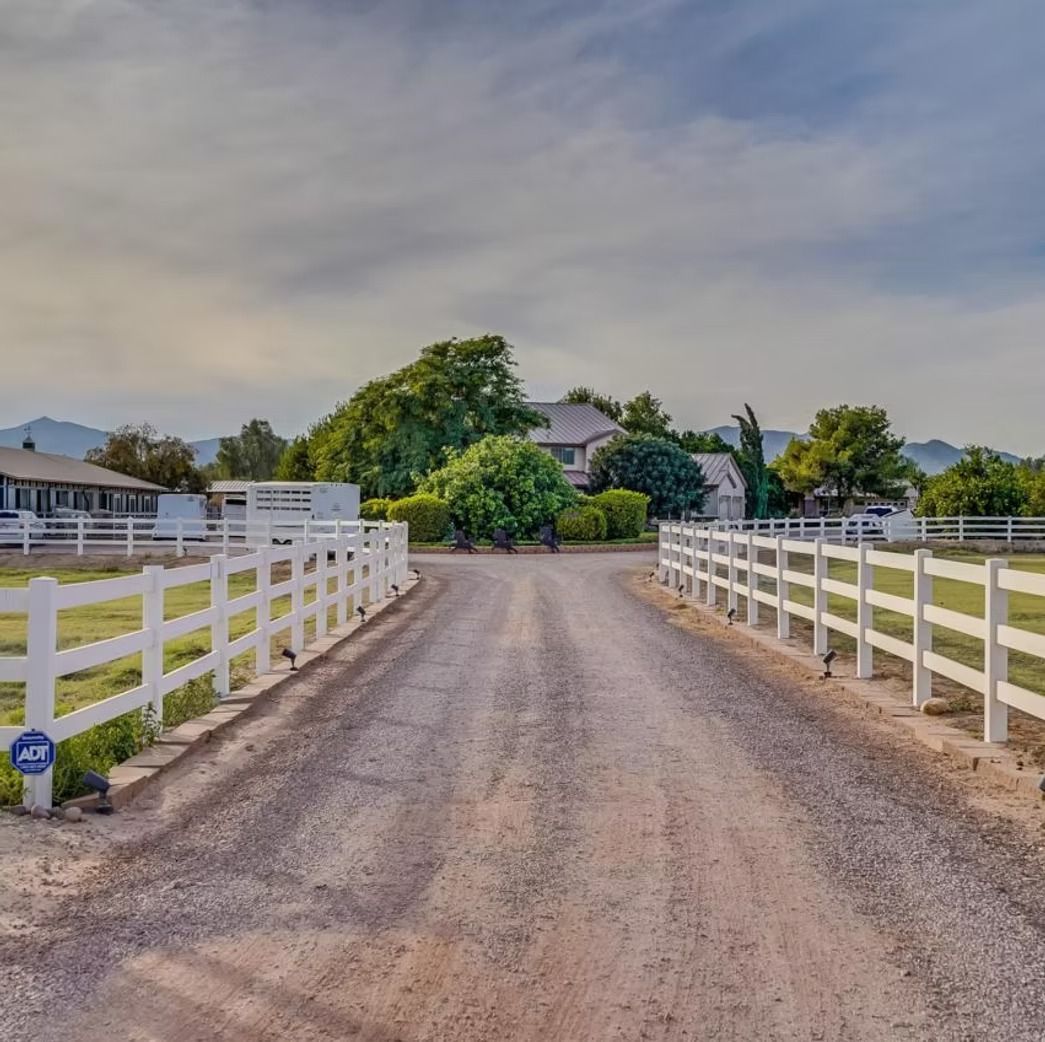 Gravel driveway between white ranch fences leading to a house with trees under a cloudy sky