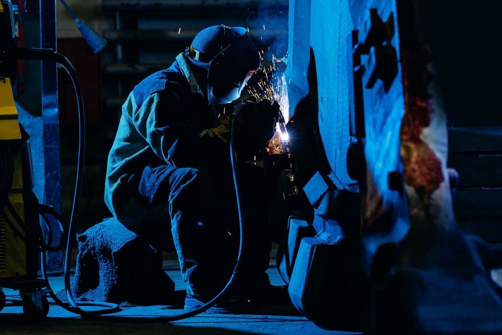 Welding worker in a blue-lit workshop, crouched beside metal machinery with bright sparks flying.