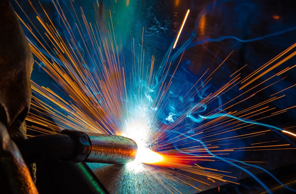Welding sparks burst from a metal pipe, with bright blue and orange light in a dark workshop.