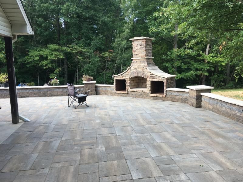 Patio with stone outdoor fireplace, paver flooring, and a chair surrounded by trees