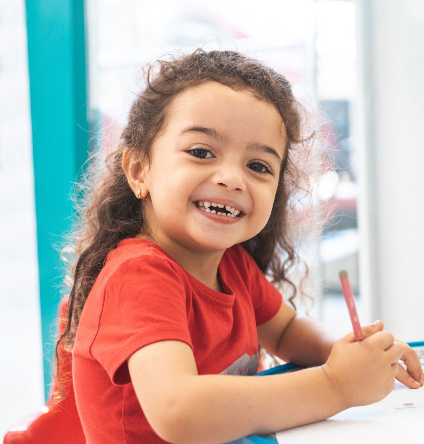 A little girl in a red shirt is sitting at a table with a pencil in her hand.