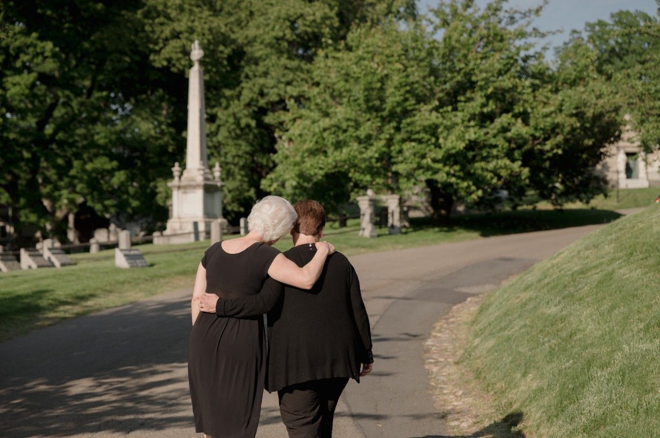 Two grieving women supporting each other while walking through a cemetery