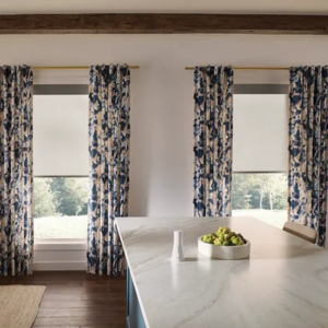 Bright kitchen with white marble island, floral curtains, and a bowl of green fruit near windows.