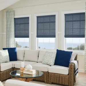 Bright sunroom with a wicker sectional, blue and white pillows, round glass coffee table, and three windows with blinds
