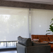 Modern living room with a dark sofa, brown chair, and large white blinds covering the windows.