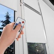 Hand holding a remote to close white window blinds beside a bright window